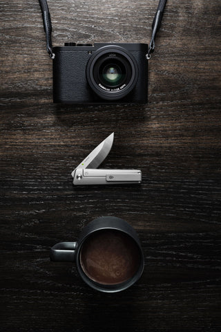 The Chapter 2 EDC frame lock titanium folding pocket knife with a camera and cup of coffee.