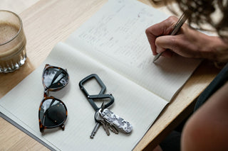 A woman sketching in her notebook while her Elko EDC keychain folding pocket knife with eco acetate scales rest a table.