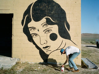 Artist Jess Mudgett painting a four eyed woman on a wall in the desert.