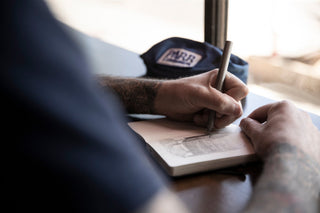 A man sketching in his notebook with the Stilwell compact travel pen.
