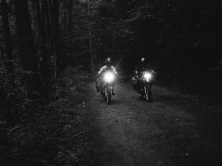 Two motorcyclist riding in the dark down a dirt road in the wilderness.